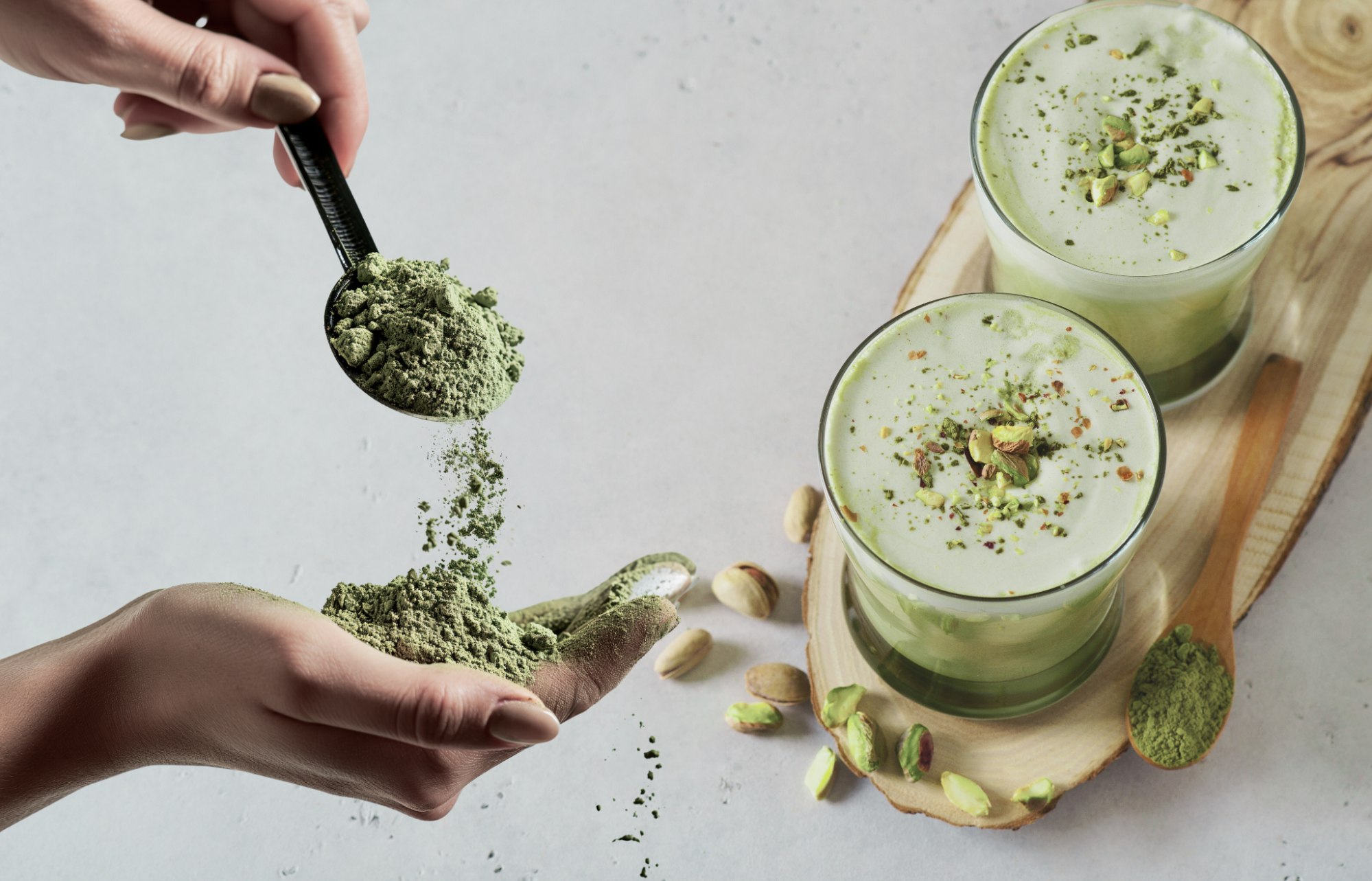 On the kitchen counter sit two glasses of green smoothie, while a hand holds a spoonful of greens powder.