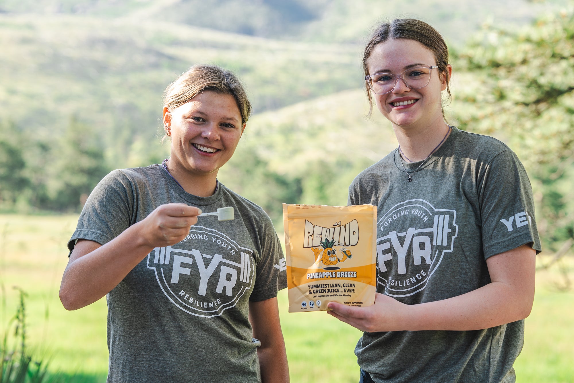 Two young women at camp FYR are smiling, holding a bag of Rewind super greens powder in a field