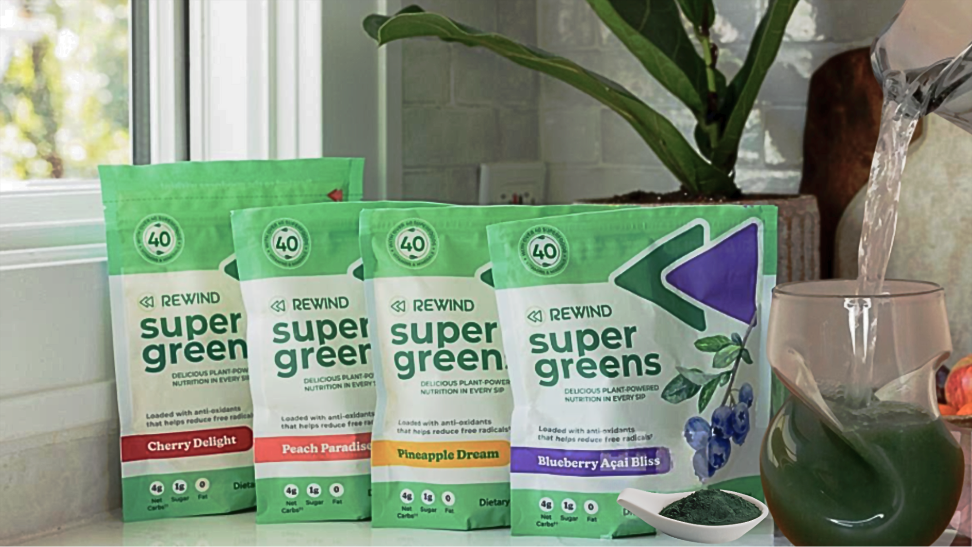 The four flavors of Rewind Greens supergreens powder are lined up on a kitchen counter with a pitcher pouring water into a glass filled with the green powder.