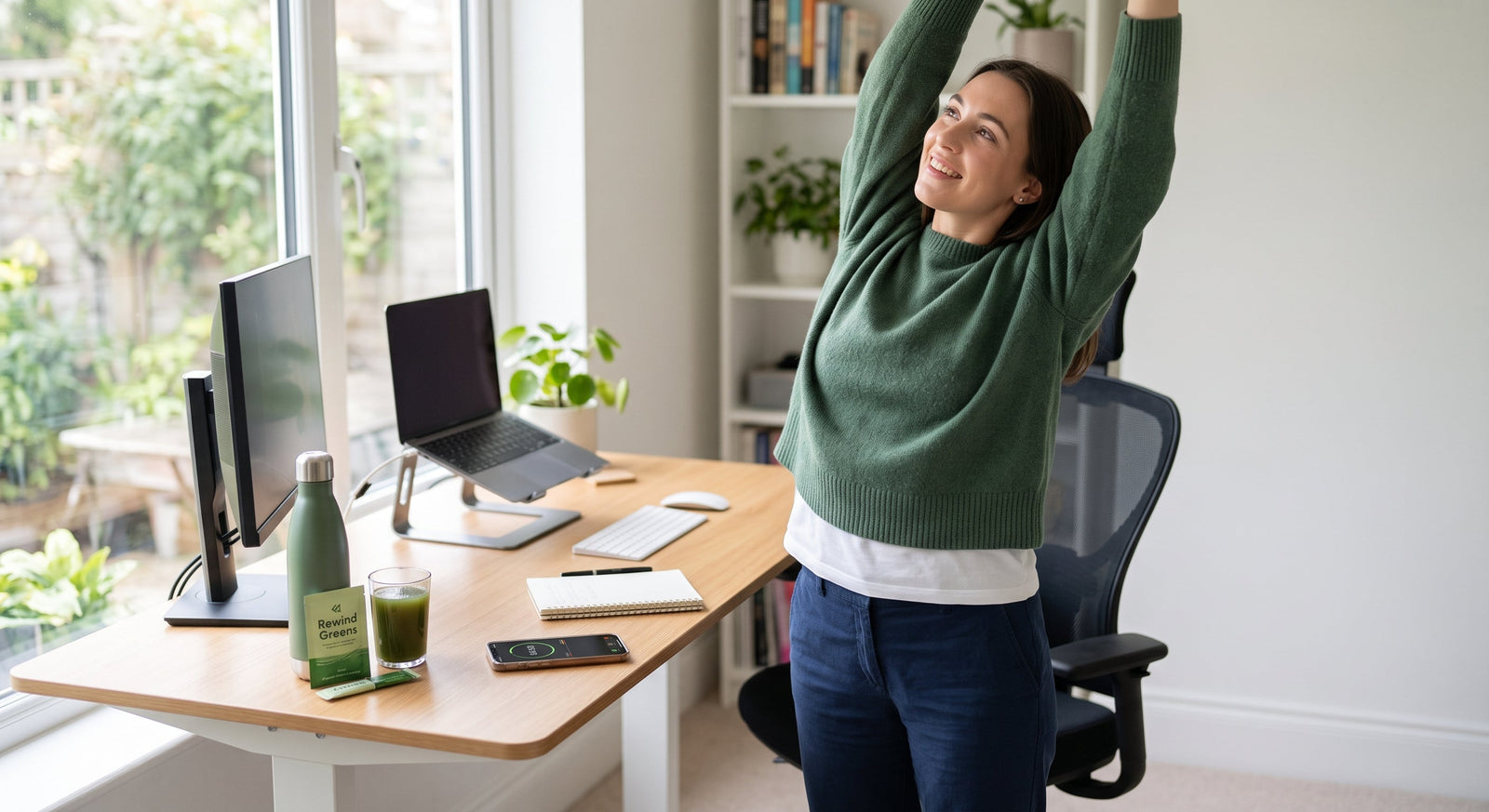 An office worker stands at her ergonomic desk taking a refreshing stretch next to a glass of green juice and a water bottle in a bright, modern workspace.